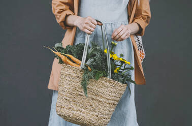 Woman holding wicker bag with fresh vegetables and flowers against gray background - NDEF00019