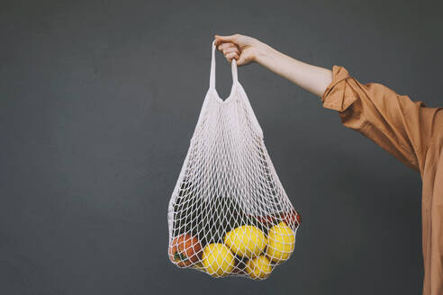 Woman holding lemons and vegetables in mesh bag against gray background - NDEF00020