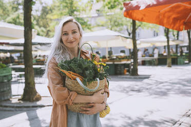 Lächelnde Frau hält eine Tasche mit Gemüse und Blumen auf dem Markt - NDEF00025