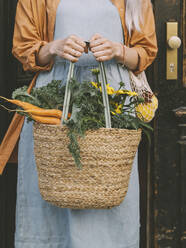 Frau hält Tasche mit Gemüse und Blumen vor einer Tür stehend - NDEF00028
