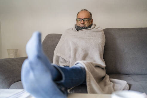 Älterer Mann, in eine Decke eingewickelt, sitzt zu Hause auf dem Sofa - DIGF19034