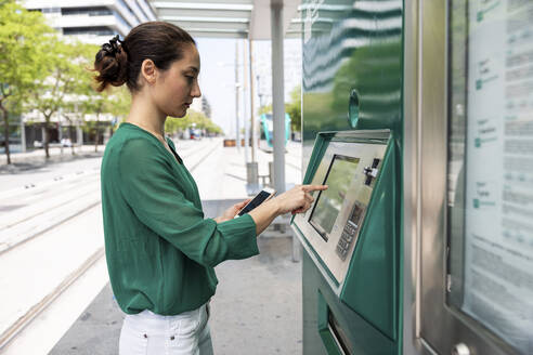 Frau mit Smartphone benutzt Fahrkartenautomat an Straßenbahnhaltestelle - WPEF06530