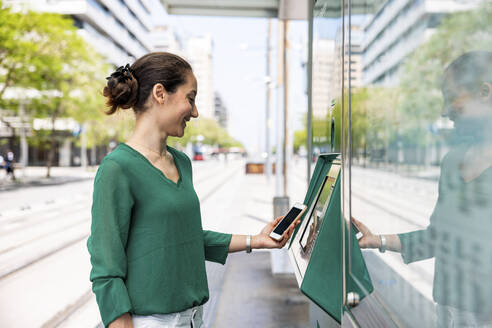 Glückliche Frau mit Smartphone und Fahrkartenautomat an einer Straßenbahnhaltestelle - WPEF06531