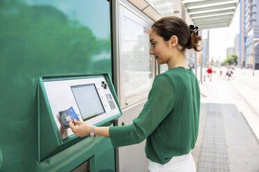 Lächelnde Frau, die einen Fahrkartenautomaten mit Kreditkarte an einer Straßenbahnhaltestelle bedient - WPEF06533