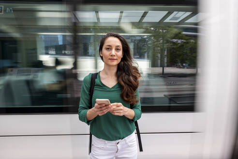 Lächelnde Frau mit Smartphone an der Straßenbahnhaltestelle - WPEF06540