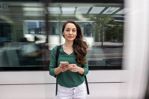 Smiling woman standing with smart phone at tram station - WPEF06540