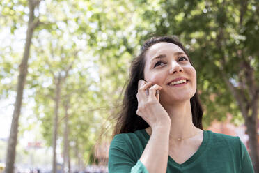 Glückliche Frau, die vor einem Baum mit ihrem Smartphone telefoniert - WPEF06552