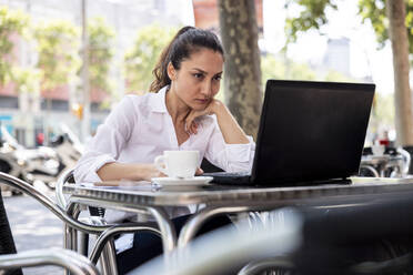 Businesswoman with hand on chin working on laptop at sidewalk cafe - WPEF06567
