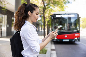 Businesswoman using smart phone standing in front of bus - WPEF06584