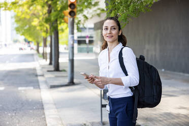 Happy businesswoman with smart phone and backpack standing at footpath - WPEF06587