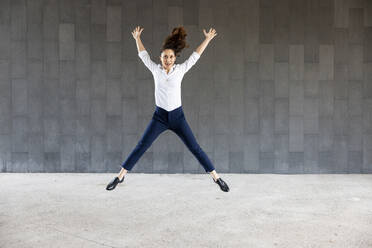 Carefree woman with arms raised jumping in front of wall - WPEF06591