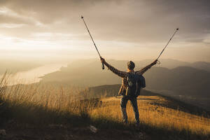 Carefree mature man holding hiking poles standing with arms raised at sunrise - UUF27587