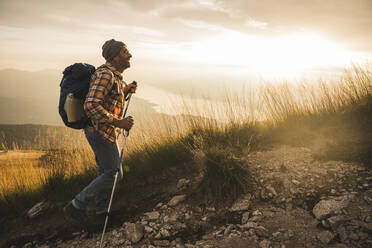 Glücklicher reifer Mann mit Rucksack beim Wandern bei Sonnenaufgang - UUF27591