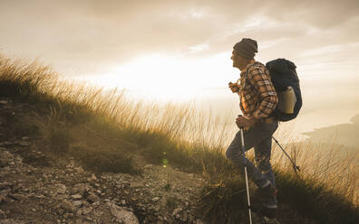 Älterer Mann mit Wanderstock und Rucksack bei Sonnenaufgang - UUF27592