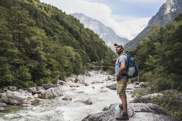 Mature man with hand on hip standing by river in front of mountains - UUF27597