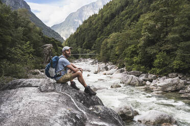 Contemplative mature man wearing cap sitting on rock by river - UUF27600