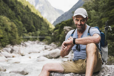 Smiling man checking time on wristwatch sitting in front of mountains - UUF27603