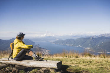 Mature woman wearing cap sitting on bench and looking at mountains - UUF27631