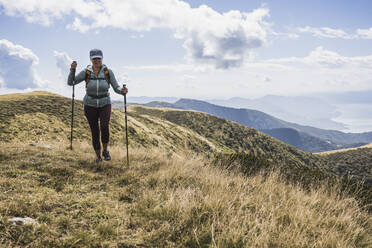 Ältere Frau geht mit Wanderstöcken auf Gras unter bewölktem Himmel - UUF27649