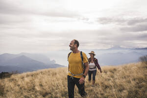 Happy mature man and woman hiking on mountain under cloudy sky - UUF27669