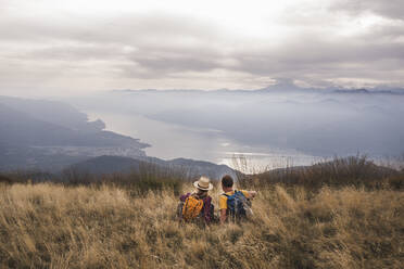 Happy mature couple sitting on grass in front of mountains under cloudy sky - UUF27680