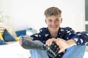 Smiling boy with prosthetic arm sitting by furniture - JOSEF14520