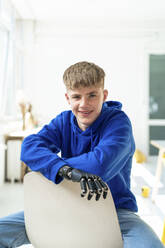 Lächelnder Jugendlicher mit amputiertem Arm, der auf einem Stuhl sitzt - JOSEF14525