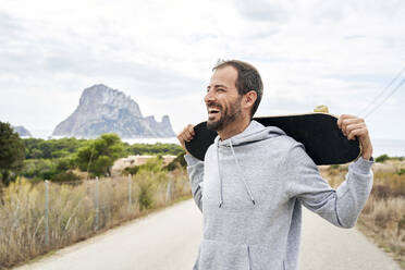 Cheerful man carrying skateboard on road - VEGF06015