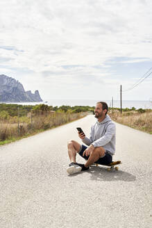 Glücklicher Mann mit Smartphone auf Skateboard sitzend - VEGF06016