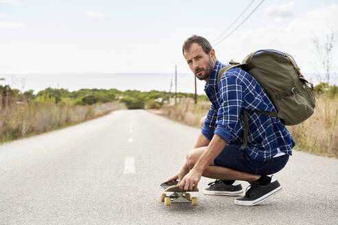 Rucksacktourist mit Skateboard auf der Straße hockend - VEGF06029