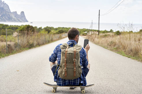 Mann mit Rucksack und Smartphone auf dem Skateboard sitzend - VEGF06032