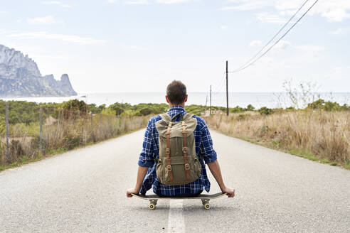 Mann mit Rucksack sitzt auf Skateboard vor Himmel - VEGF06034