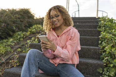 Smiling blond woman using smart phone on staircase - JCCMF07629