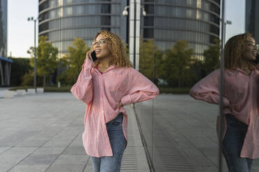 Smiling woman with hand on hip talking on smart phone by glass wall - JCCMF07634