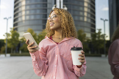 Lächelnde Frau mit Mobiltelefon und Einweg-Kaffeebecher an einer Glaswand - JCCMF07639