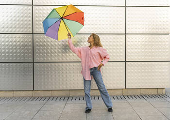 Junge Frau spielt mit buntem Regenschirm vor einer Mauer - JCCMF07640