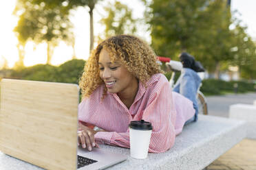 Smiling young woman with disposable coffee cup using laptop on bench - JCCMF07645