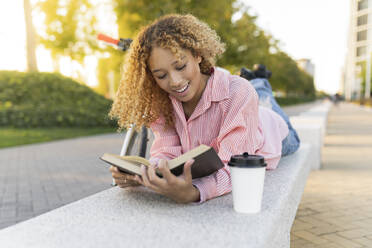 Smiling woman reading book lying on bench - JCCMF07646