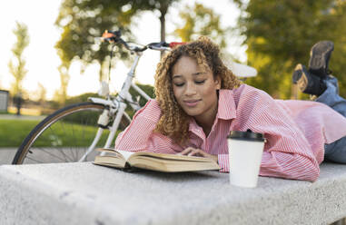 Young woman with hand in hair reading book on bench - JCCMF07647