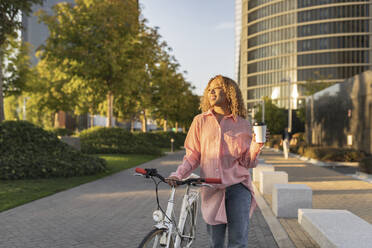 Smiling woman holding disposable coffee cup and walking with bicycle on footpath - JCCMF07653