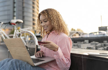 Lächelnde Frau mit Kreditkarte beim Online-Shopping auf einem Laptop an einer Bank - JCCMF07660