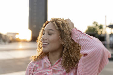 Smiling young woman with hand in hair and eyes closed at sunset - JCCMF07664