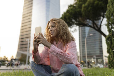 Lächelnde Frau mit Smartphone, die auf einer Wiese in der Stadt nachdenkt - JCCMF07666