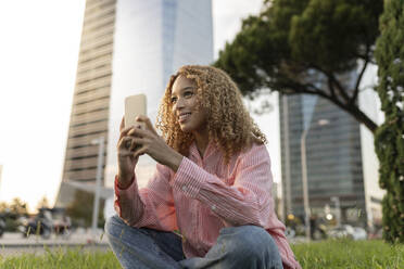 Smiling woman with smart phone contemplating on grass in city - JCCMF07666