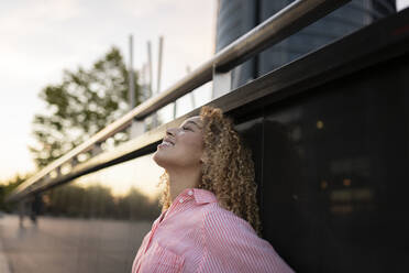 Smiling woman day dreaming with eyes closed in front of wall - JCCMF07676