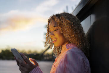 Junge lockig behaarte Frau benutzt Smartphone bei Sonnenuntergang - JCCMF07678