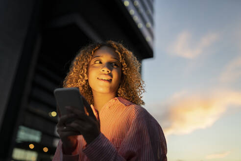 Lächelnde Frau mit Handy, die bei Sonnenuntergang nachdenkt - JCCMF07679
