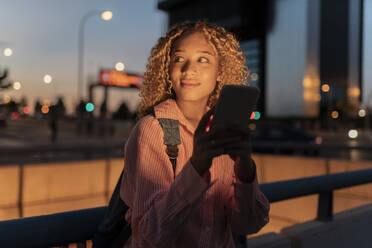 Smiling young woman with smart phone by railing at night - JCCMF07687