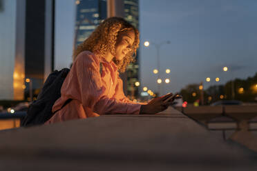 Lächelnde Frau, die sich an eine Wand lehnt und ein Mobiltelefon in der Stadt benutzt - JCCMF07689