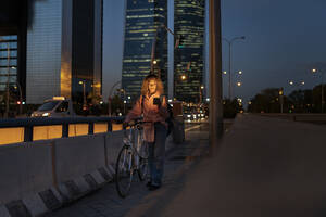 Junge Frau, die ein Mobiltelefon benutzt und mit dem Fahrrad auf einer Straße in der Stadt spazieren geht - JCCMF07690